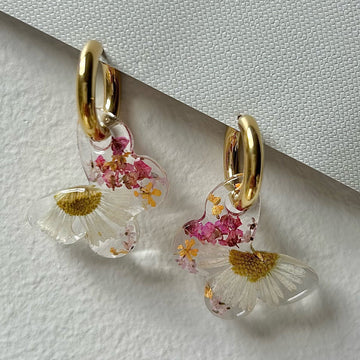 A pair of floral resin earrings with real forget-me-nots in gold bezels, hanging from gold huggie hoops, displayed on a clean cream-toned background with soft light.