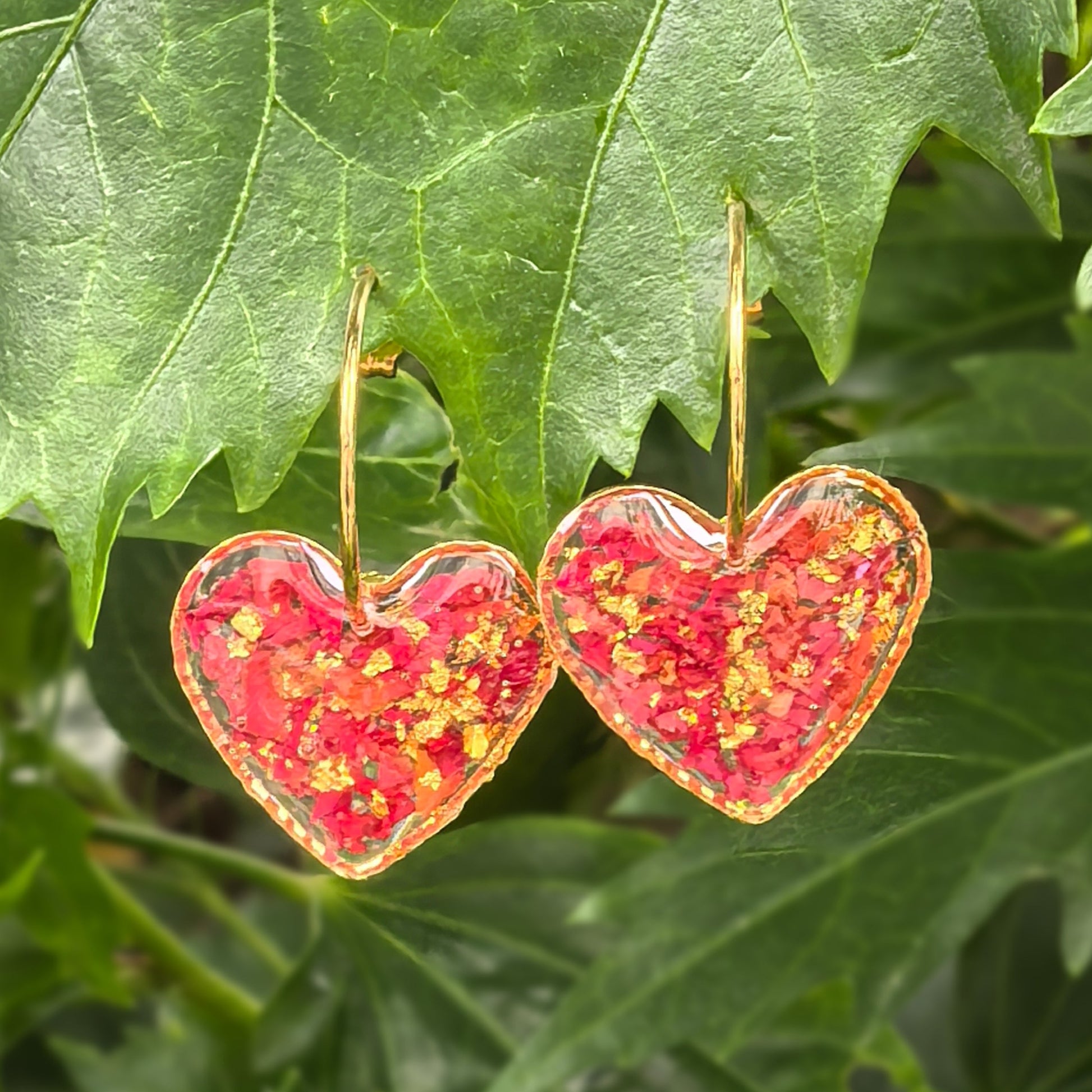 Heart-shaped earrings encapsulating red rose petals, hanging on green leaves.