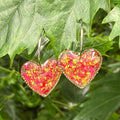 Heart-shaped earrings encapsulating red rose petals, hanging on green leaves.