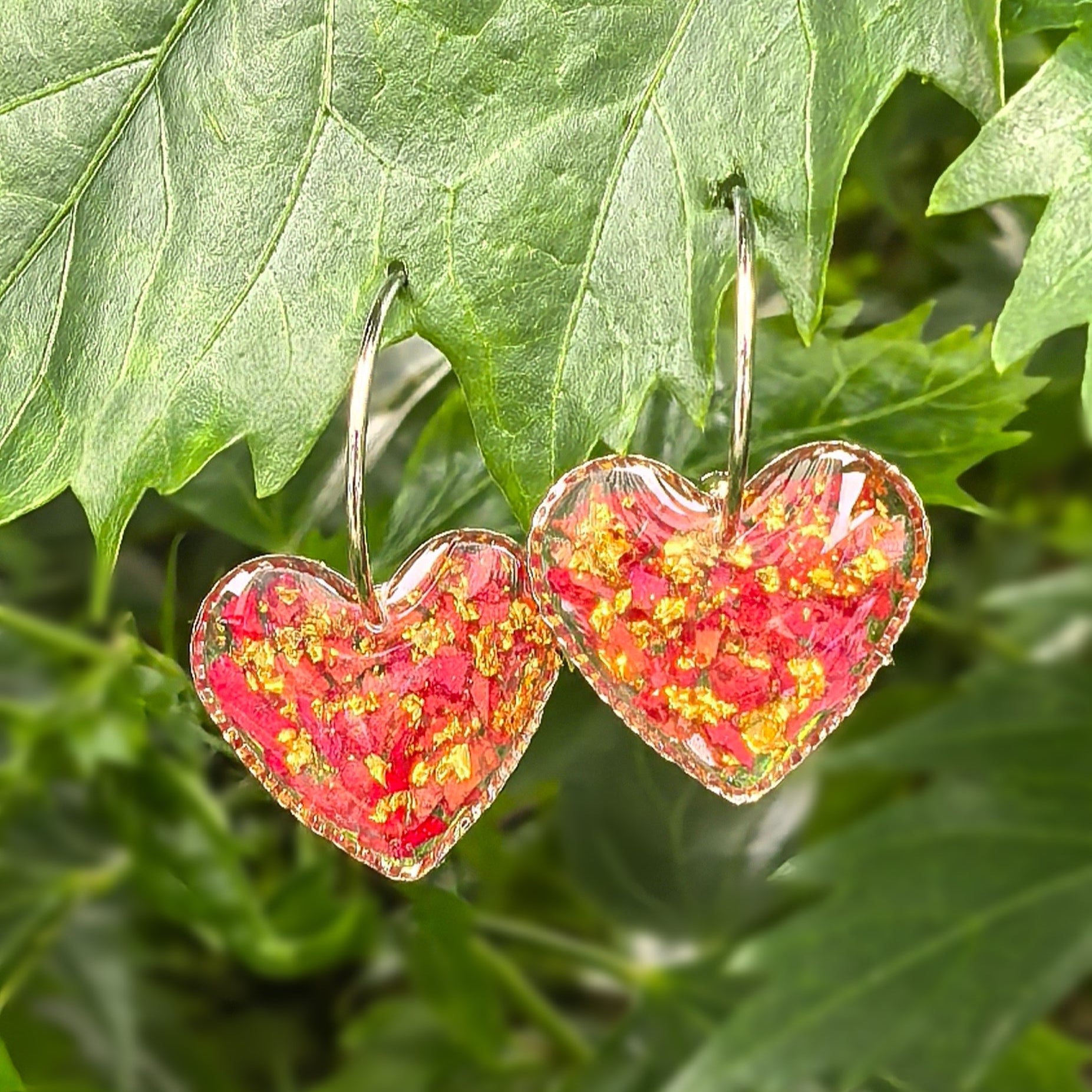 Heart-shaped earrings encapsulating red rose petals, hanging on green leaves.