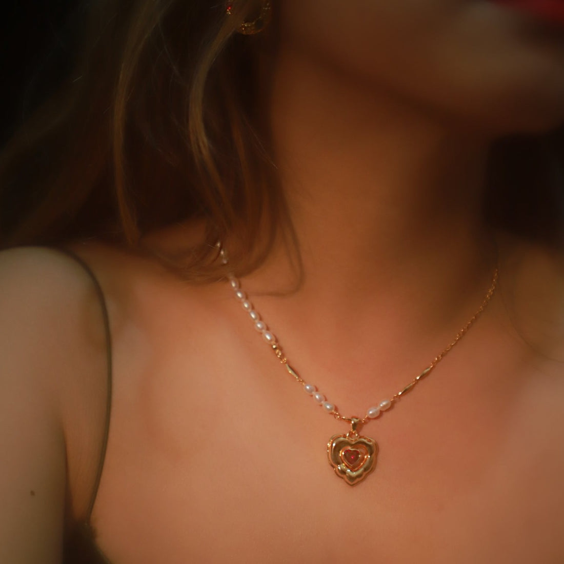 A blonde woman wearing the Crimson Love necklace, featuring a heart-shaped red crystal and freshwater pearls.