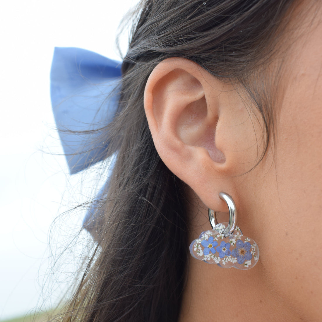 Close-up of cloud-shaped earrings filled with real dried forget-me-nots,  worn on a woman's ear.