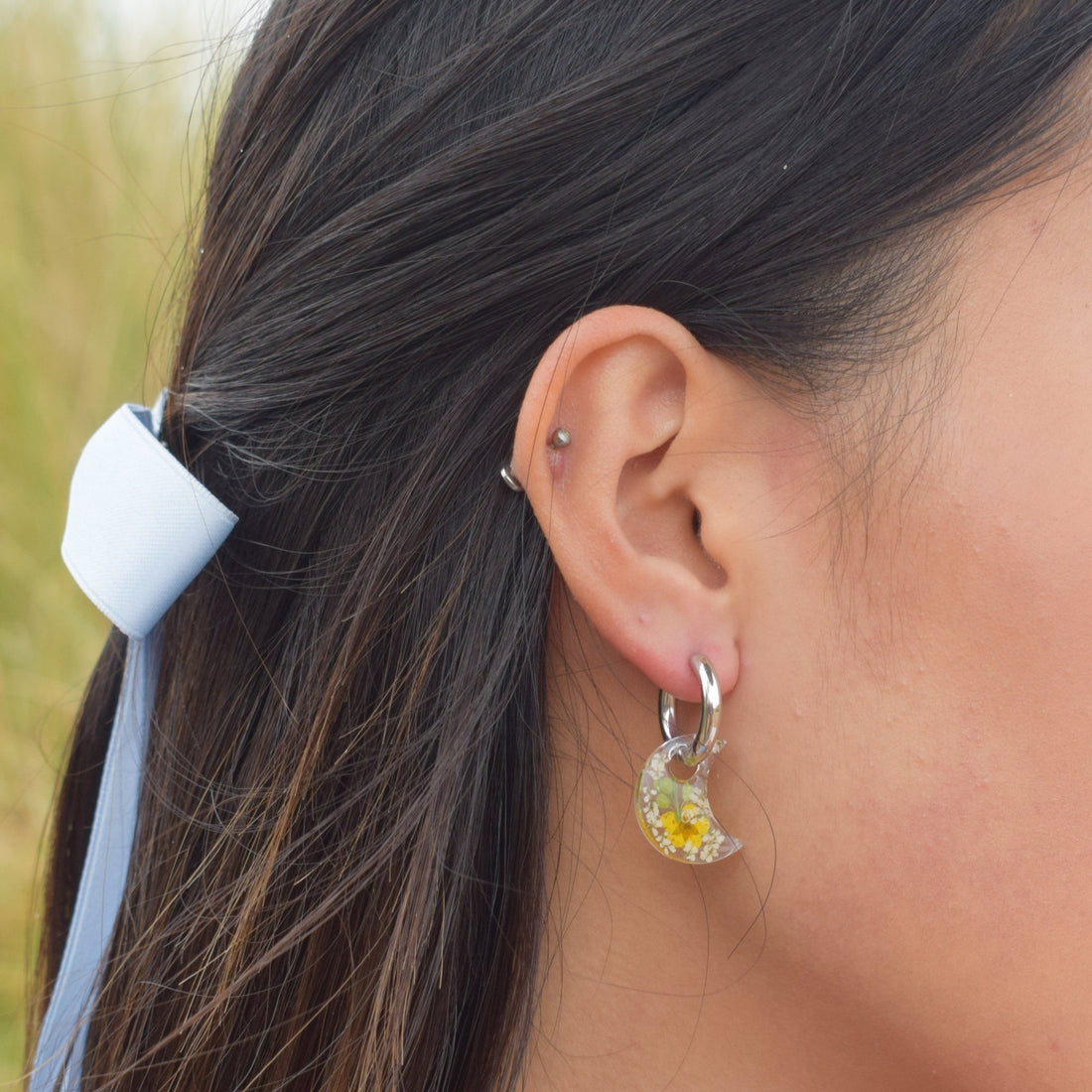 A pair of crescent huggie earrings with real flowers, worn on a woman's ear.