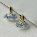 Close-up of cloud-shaped earrings filled with real dried forget-me-nots, hanging from gold huggie hoops, styled on a cream-toned studio background with bright soft lighting.