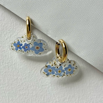 Close-up of cloud-shaped earrings filled with real dried forget-me-nots, hanging from gold huggie hoops, styled on a cream-toned studio background with bright soft lighting.