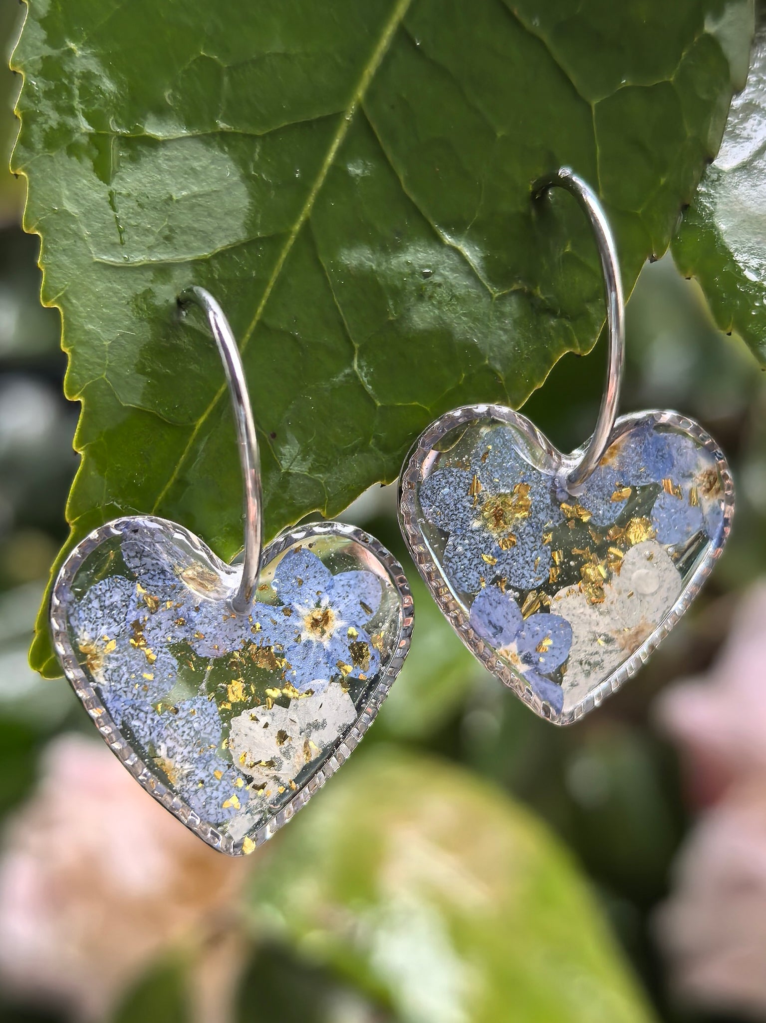 A pair of heart-shaped earrings made with forget-me-not flowers and stainless steel hoops, hanging gracefully on green leaves.