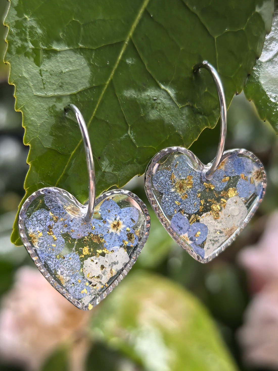 A pair of heart-shaped earrings made with forget-me-not flowers and stainless steel hoops, hanging gracefully on green leaves.