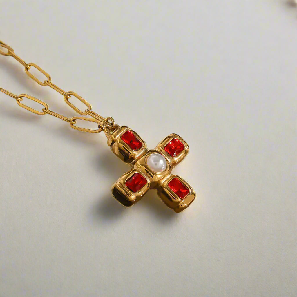 Alt text: A gold-plated stainless steel cross necklace featuring deep red cubic zirconia stones at each end and a central faux pearl, set against a clean, bright background.