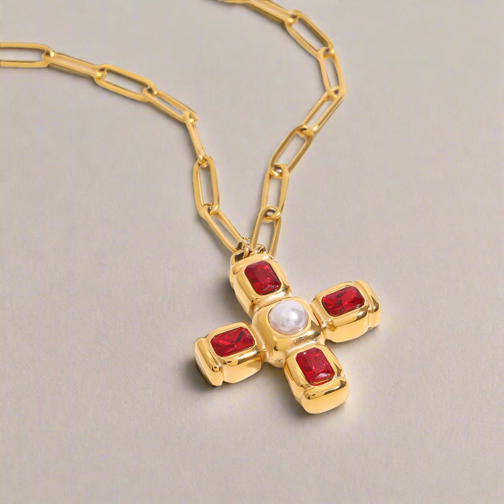 A gold-plated stainless steel cross necklace featuring deep red cubic zirconia stones at each end and a central faux pearl, set against a clean, bright background.